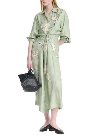 A woman wears the Floyd Dress by Altuzarra—a light green, long-sleeve shirt dress with a point collar, button-down front, abstract print, and wrap tie waist—paired with a black handbag and black flats.