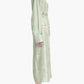 A person stands in profile wearing the Altuzarra Floyd Dress—a long, light green shirt dress with floral embroidery, a point collar—and black flat shoes.