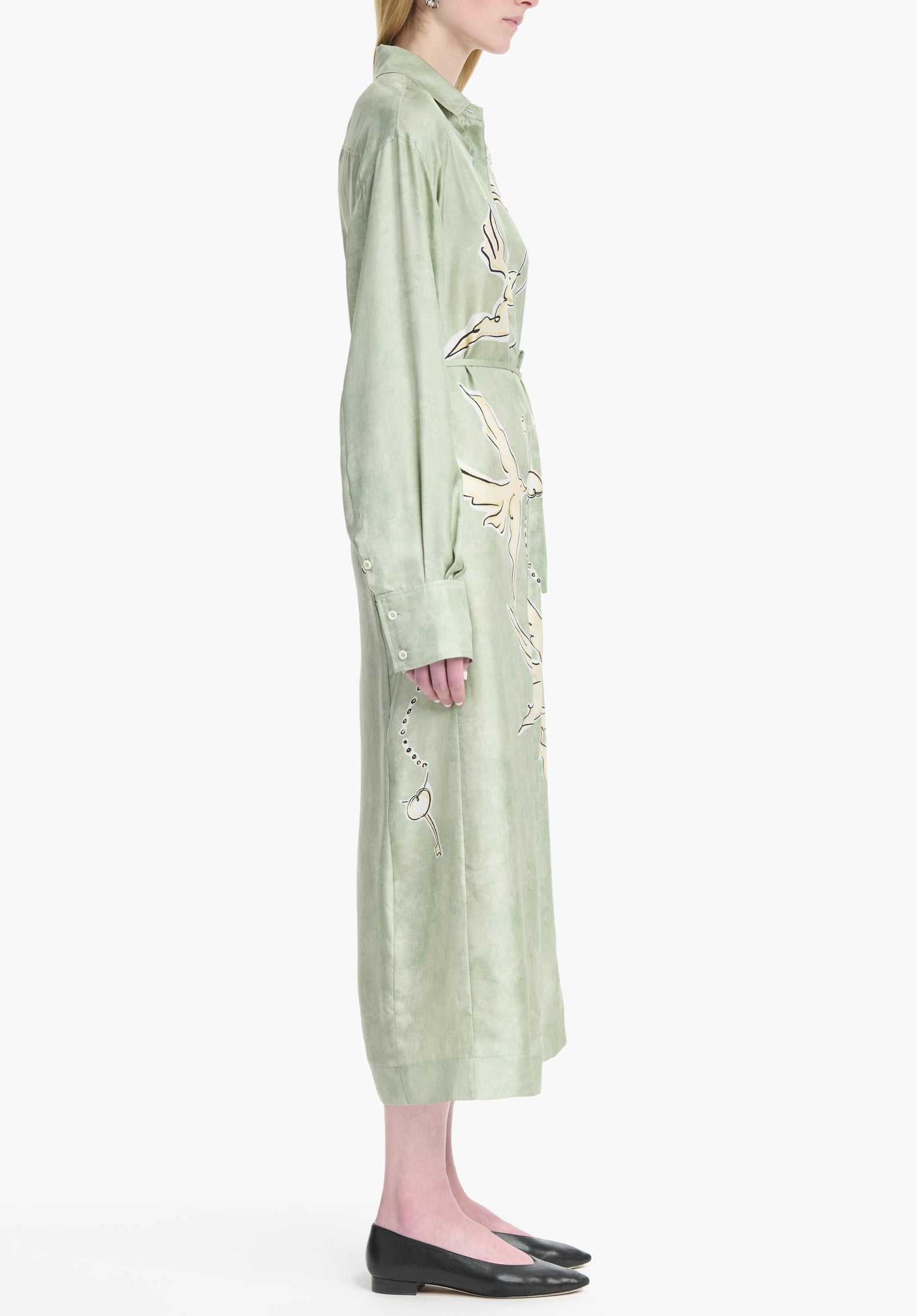 A person stands in profile wearing the Altuzarra Floyd Dress—a long, light green shirt dress with floral embroidery, a point collar—and black flat shoes.