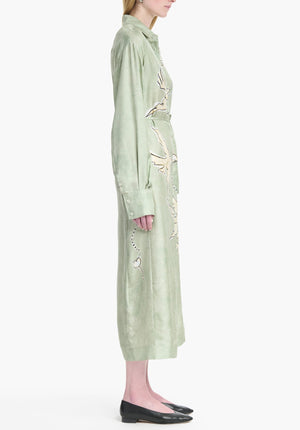 A person stands in profile wearing the Altuzarra Floyd Dress—a long, light green shirt dress with floral embroidery, a point collar—and black flat shoes.