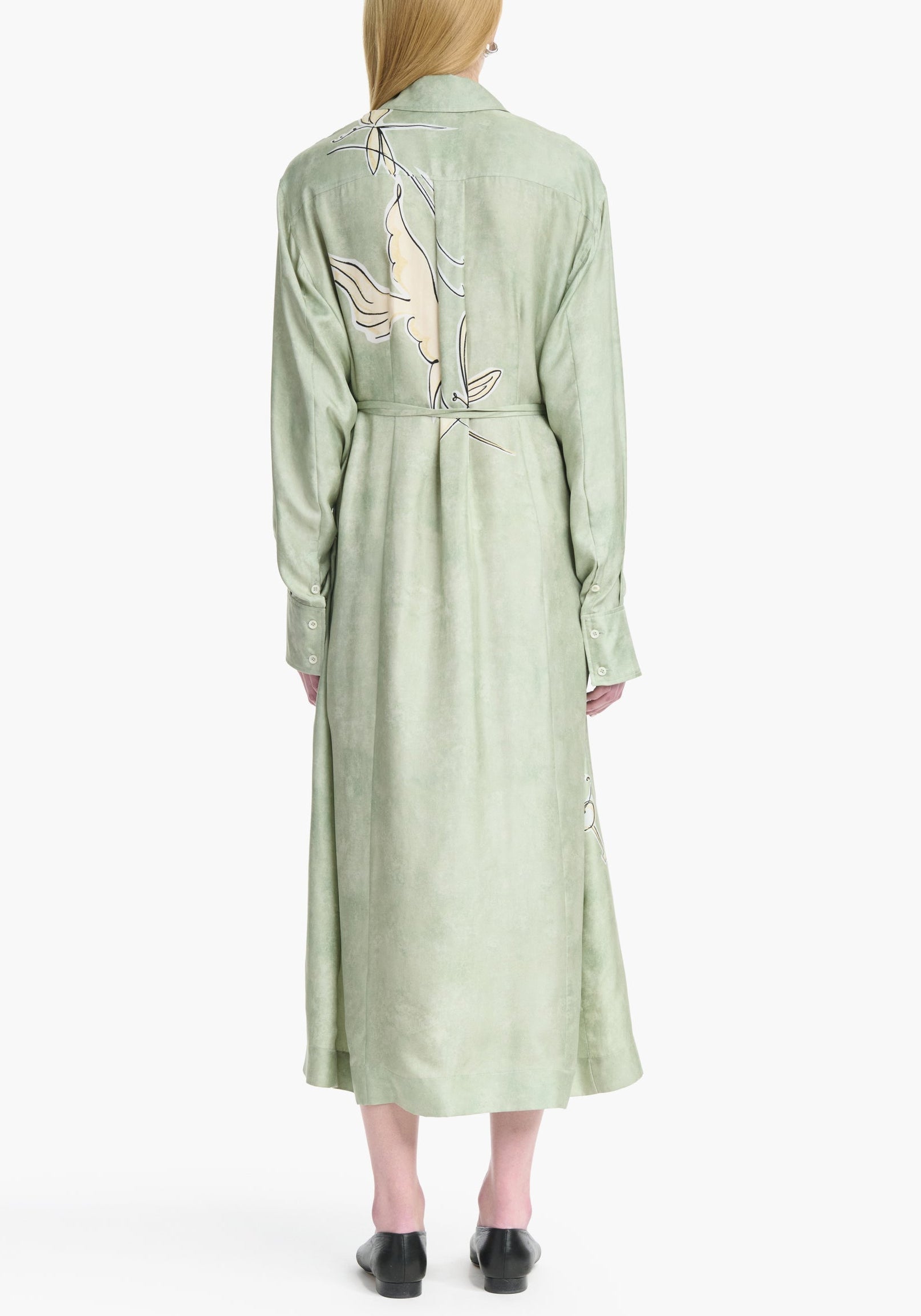 A woman stands with her back to the camera, wearing the Altuzarra Floyd Dress—a light green, long-sleeved shirt dress featuring a classic point collar and a subtle abstract pattern on the back.