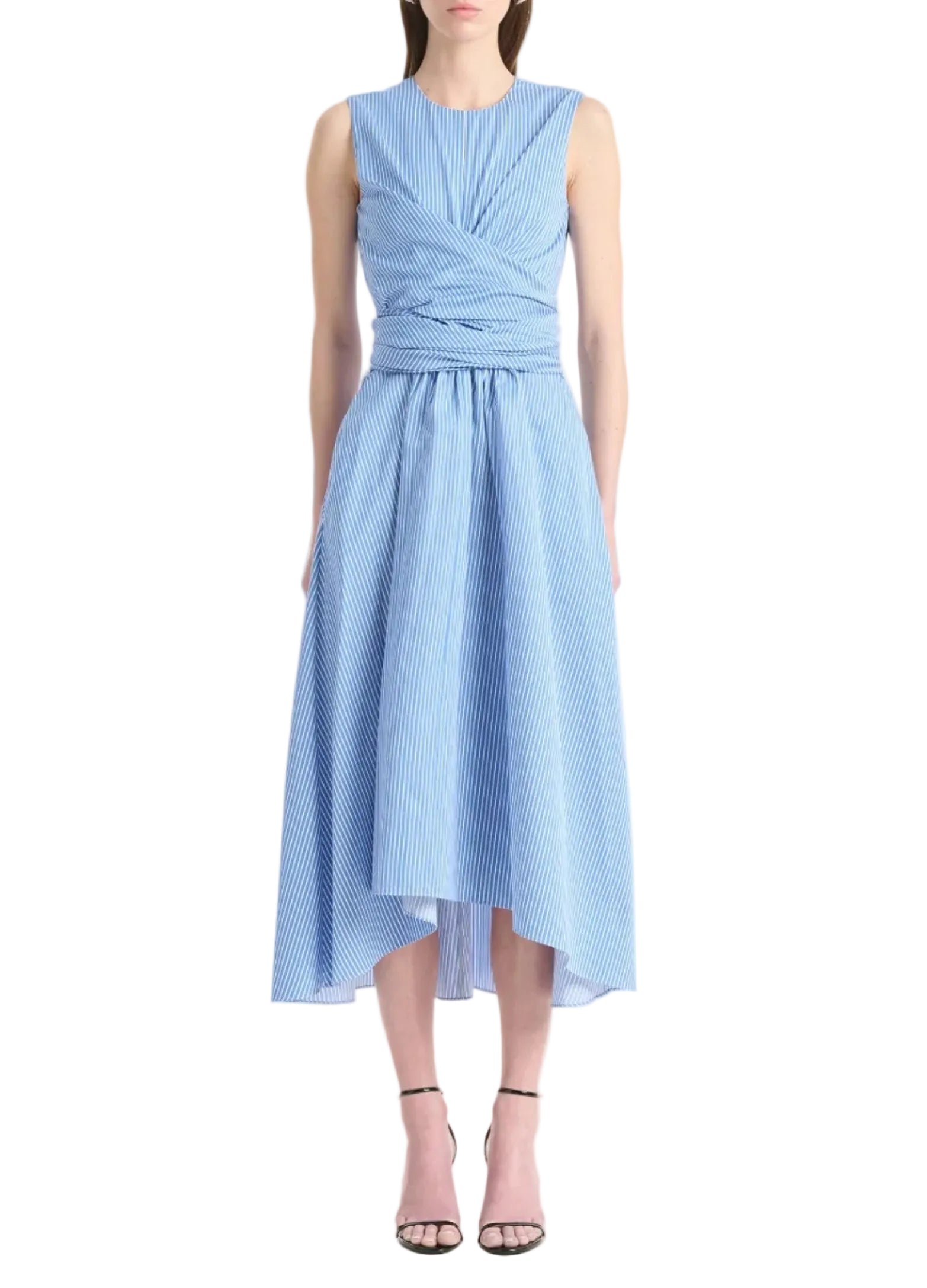 A woman wears the Altuzarra Penny Dress—a sleeveless cotton dress with blue and white stripes, fitted bodice, wrap waist, and asymmetric high-low hem—paired with black ankle-strap heels.