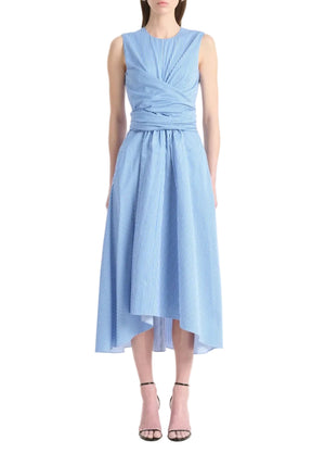 A woman wears the Altuzarra Penny Dress—a sleeveless cotton dress with blue and white stripes, fitted bodice, wrap waist, and asymmetric high-low hem—paired with black ankle-strap heels.