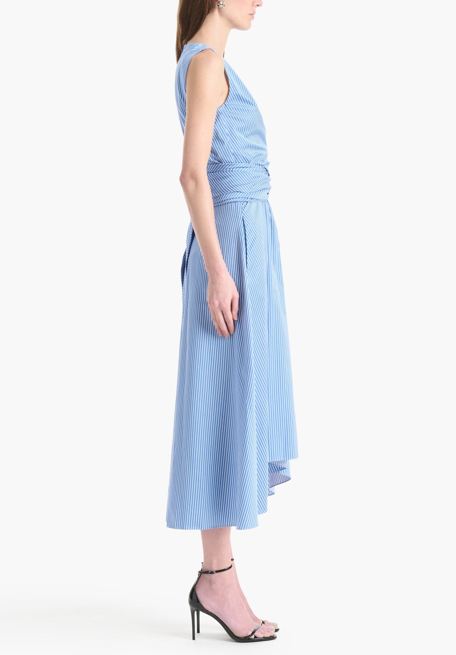 A woman stands in profile wearing the Altuzarra Penny Dress, a sleeveless cotton design with blue and white stripes, wrapped waist, asymmetric hem, and paired with black high-heeled sandals.