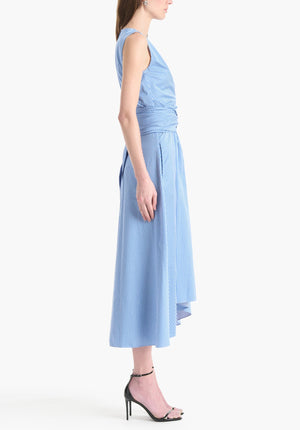 A woman stands in profile wearing the Altuzarra Penny Dress, a sleeveless cotton design with blue and white stripes, wrapped waist, asymmetric hem, and paired with black high-heeled sandals.