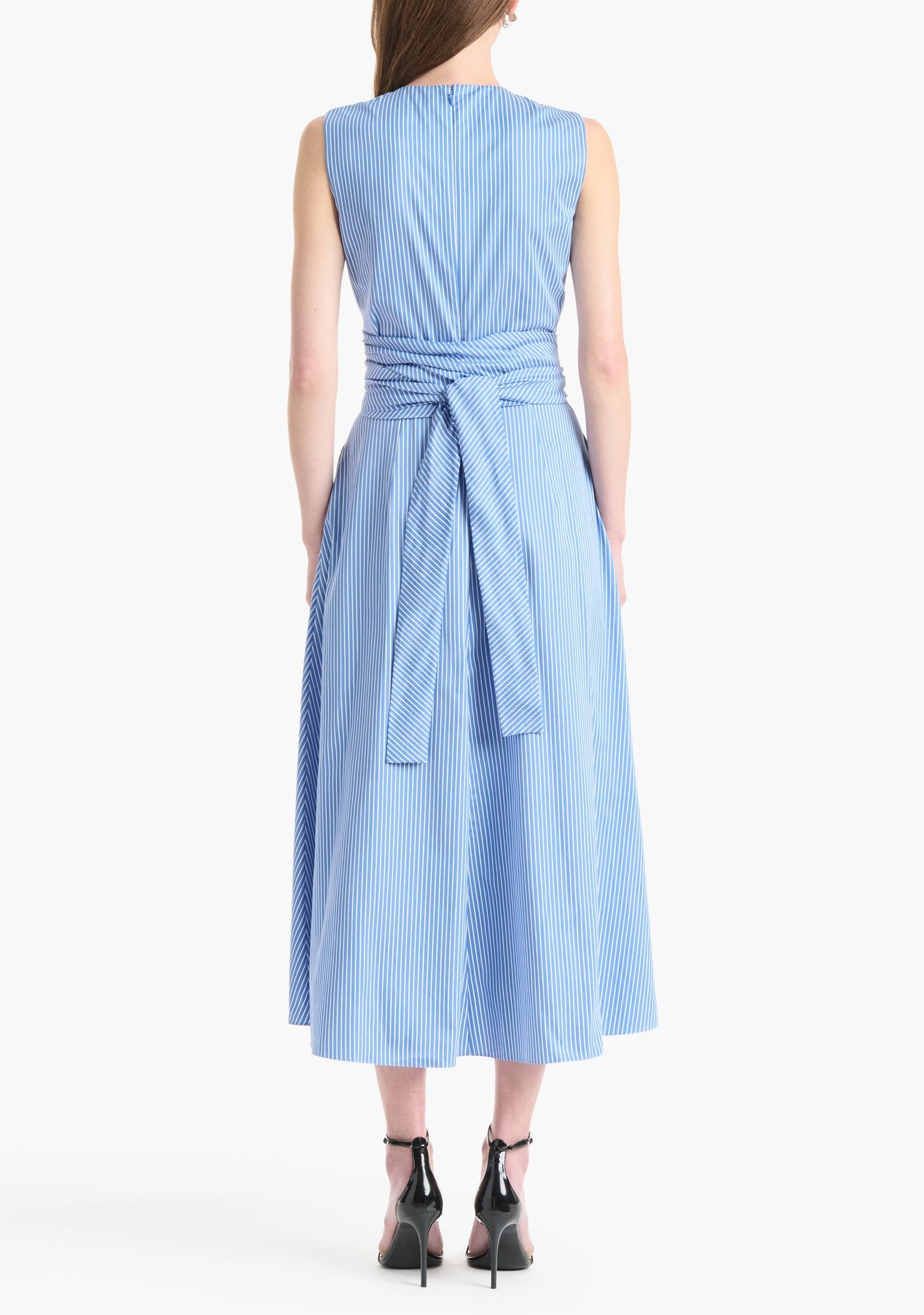 A woman shown from the back wears the Altuzarra Penny Dress, a sleeveless cotton design with blue and white stripes, a tied waist belt, and an asymmetric hem, paired with black heeled shoes.