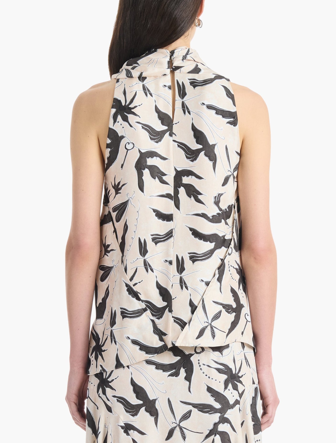 A woman is seen from behind wearing the Altuzarra Sleary Top, a sleeveless cream silk piece featuring a black bird and foliage print, paired with a matching skirt.