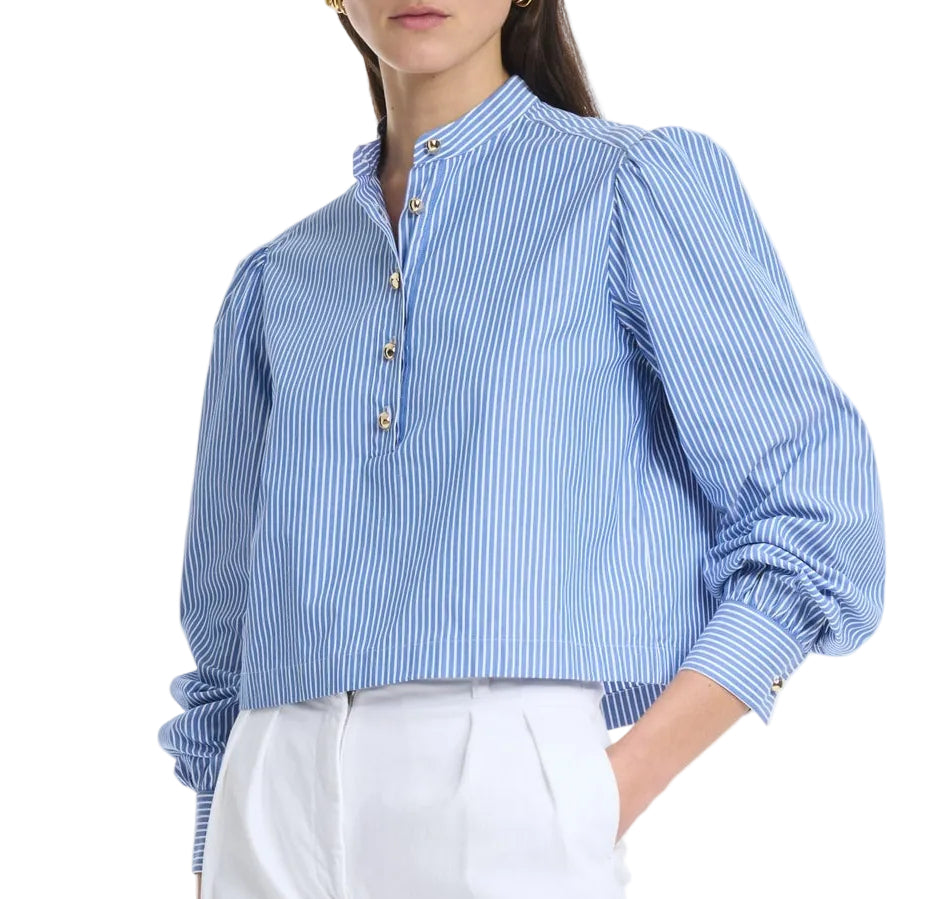 A woman wears the Altuzarra Margot Top—a blue and white vertically striped cotton-poplin blouse with gold buttons and subtle balloon sleeves—paired with high-waisted white trousers.