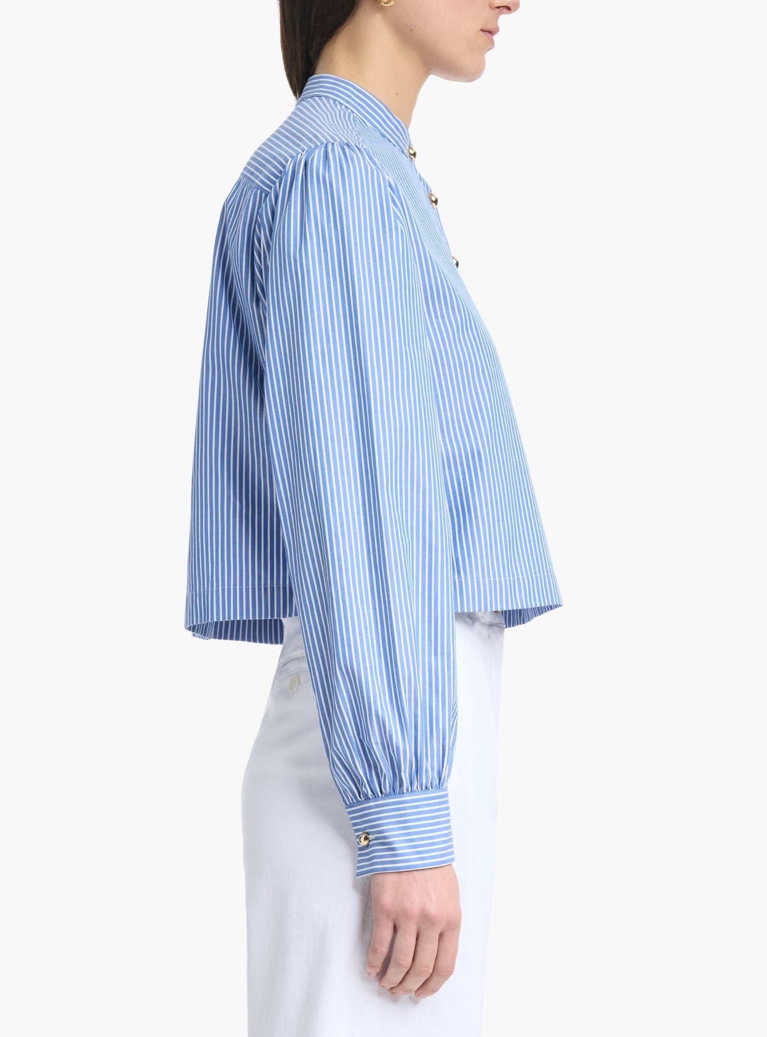 A woman is shown from the side against a plain background, wearing the Altuzarra Margot Top—a blue and white vertical striped modern shirt with balloon sleeves and gold buttons—paired with white high-waisted pants.