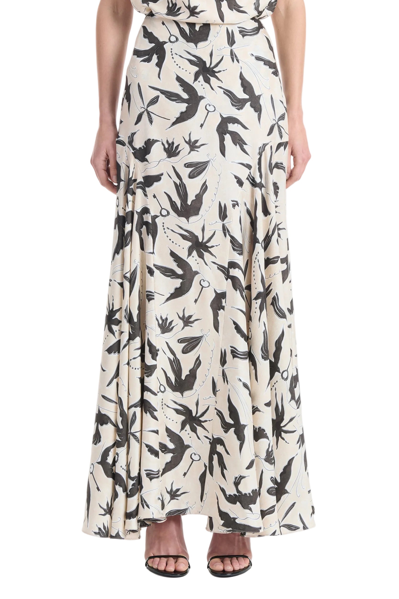 A person wearing the Altuzarra Elton Skirt—a cream, ankle-length skirt with a black bird print and flared silhouette—paired with open-toed black heels.