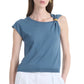 A woman stands against a plain background wearing the Altuzarra Rinaldi Top—a blue jersey knit style featuring gold metal hardware on one shoulder—paired with white pants.