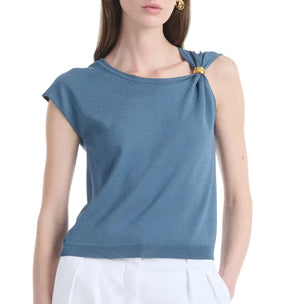 A woman stands against a plain background wearing the Altuzarra Rinaldi Top—a blue jersey knit style featuring gold metal hardware on one shoulder—paired with white pants.