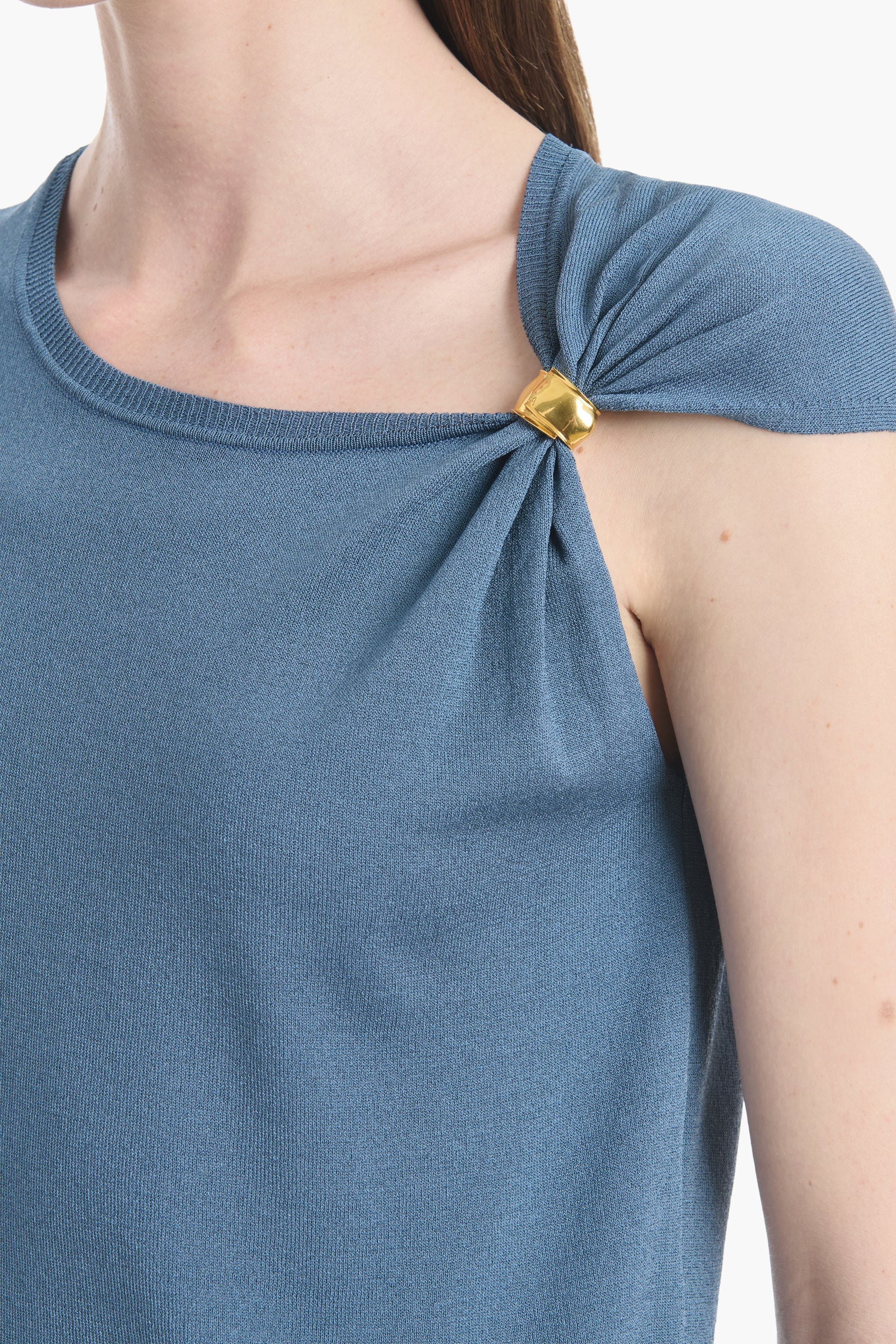A close-up of a person wearing the Altuzarra Rinaldi Top, a blue jersey knit style with gathered shoulders accented by gold metal details.