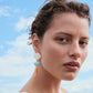 A person with short, wavy hair wears a white top and bright Luna Drop Earrings by Lizzie Fortunato, featuring amazonite beads, while standing outdoors beneath a scattered cloud sky.
