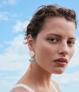 A person with short, wavy hair wears a white top and bright Luna Drop Earrings by Lizzie Fortunato, featuring amazonite beads, while standing outdoors beneath a scattered cloud sky.