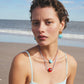 A woman with short hair wears a light blue swimsuit and Lizzie Fortunato's Kai Earrings, along with colorful shell-themed jewelry, standing on a sandy beach with the ocean and sky in the background.