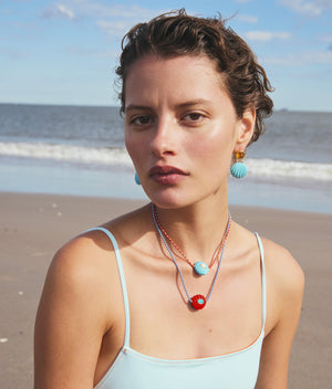 A woman with short hair wears a light blue swimsuit and Lizzie Fortunato's Kai Earrings, along with colorful shell-themed jewelry, standing on a sandy beach with the ocean and sky in the background.