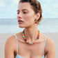 A woman with short hair in a blue swimsuit sits on the beach, the ocean and sky behind her. She wears Laurel Earrings by Lizzie Fortunato, adding a touch of color and charm to the scene with their vibrant beaded design.