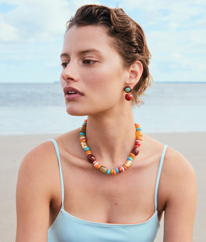 A woman with short hair in a blue swimsuit sits on the beach, the ocean and sky behind her. She wears Laurel Earrings by Lizzie Fortunato, adding a touch of color and charm to the scene with their vibrant beaded design.