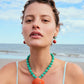 A woman wearing the Lizzie Fortunato Corfu Shell Necklace, featuring amazonite beads and a coral-toned ceramic shell pendant with matching earrings, stands on a beach with the ocean behind her.