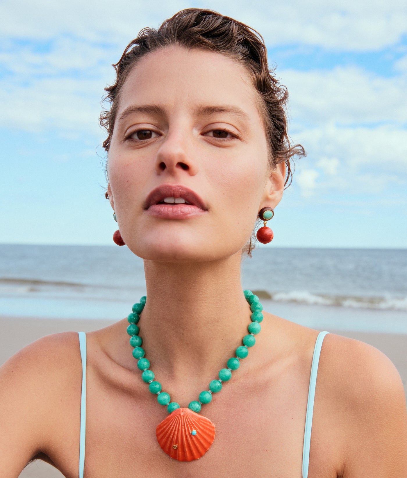 A woman wearing the Lizzie Fortunato Corfu Shell Necklace, featuring amazonite beads and a coral-toned ceramic shell pendant with matching earrings, stands on a beach with the ocean behind her.