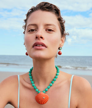 A woman wearing the Lizzie Fortunato Corfu Shell Necklace, featuring amazonite beads and a coral-toned ceramic shell pendant with matching earrings, stands on a beach with the ocean behind her.
