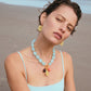 A woman in a light blue swimsuit poses on the beach, wearing the Cordoba Necklace by Lizzie Fortunato with matching earrings. The ocean and sand are visible in the background.