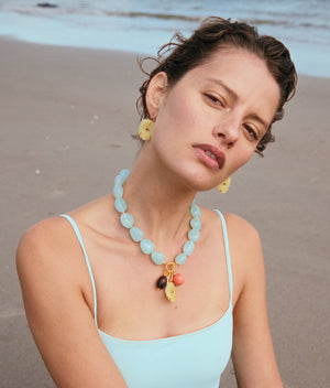 A woman in a light blue swimsuit poses on the beach, wearing the Cordoba Necklace by Lizzie Fortunato with matching earrings. The ocean and sand are visible in the background.