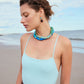 A woman in a light blue swimsuit and the Lizzie Fortunato Lita Necklace stands on a sandy beach, gazing downward with the ocean and sky behind her.