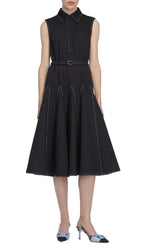 A woman wearing the elegant Monique Denim Dress by Emilia Wickstead—a sleeveless black midi dress with white stitching, featuring a belted waist and flared skirt—paired with striking blue and black heeled shoes.