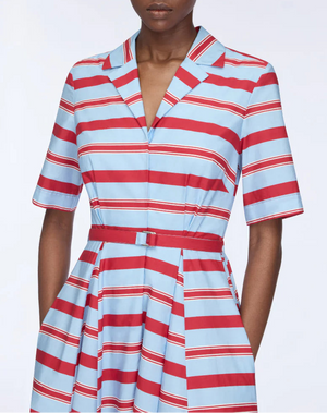 A person wears the Emilia Wickstead Mella Bayadere Stripe Dress, featuring red, white, and blue horizontal stripes, a V-neck collar, pockets, short sleeves, and a matching belt at the waist.