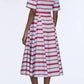 A person stands with their back to the camera, wearing the Emilia Wickstead Mella Bayadere Stripe Dress—a knee-length, short-sleeved style featuring horizontal red and aqua stripes—paired with black heeled mules.