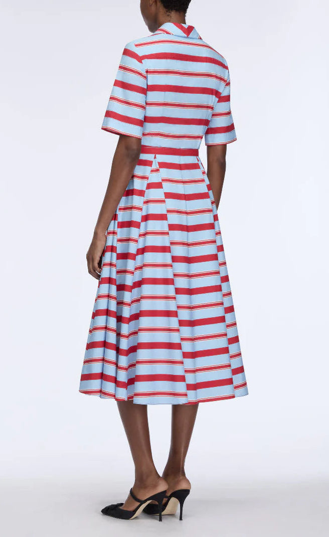 A person stands with their back to the camera, wearing the Emilia Wickstead Mella Bayadere Stripe Dress—a knee-length, short-sleeved style featuring horizontal red and aqua stripes—paired with black heeled mules.