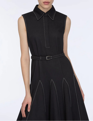 A woman wears the Emilia Wickstead Monique Denim Dress—a sleeveless black collared dress with white stitching, a belted waist, and a flared skirt.