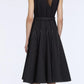 A woman stands with her back to the camera, wearing the Emilia Wickstead Monique Denim Dress—a sleeveless black dress with white stitching and a belted waist—paired with multicolored heels.