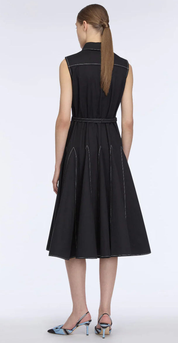 A woman stands with her back to the camera, wearing the Emilia Wickstead Monique Denim Dress—a sleeveless black dress with white stitching and a belted waist—paired with multicolored heels.