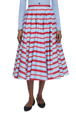 A person wearing a blue long-sleeve top and the Emilia Wickstead Rhea Skirt—a high-waisted, tea-length skirt featuring red and white horizontal stripes—paired with black flats.