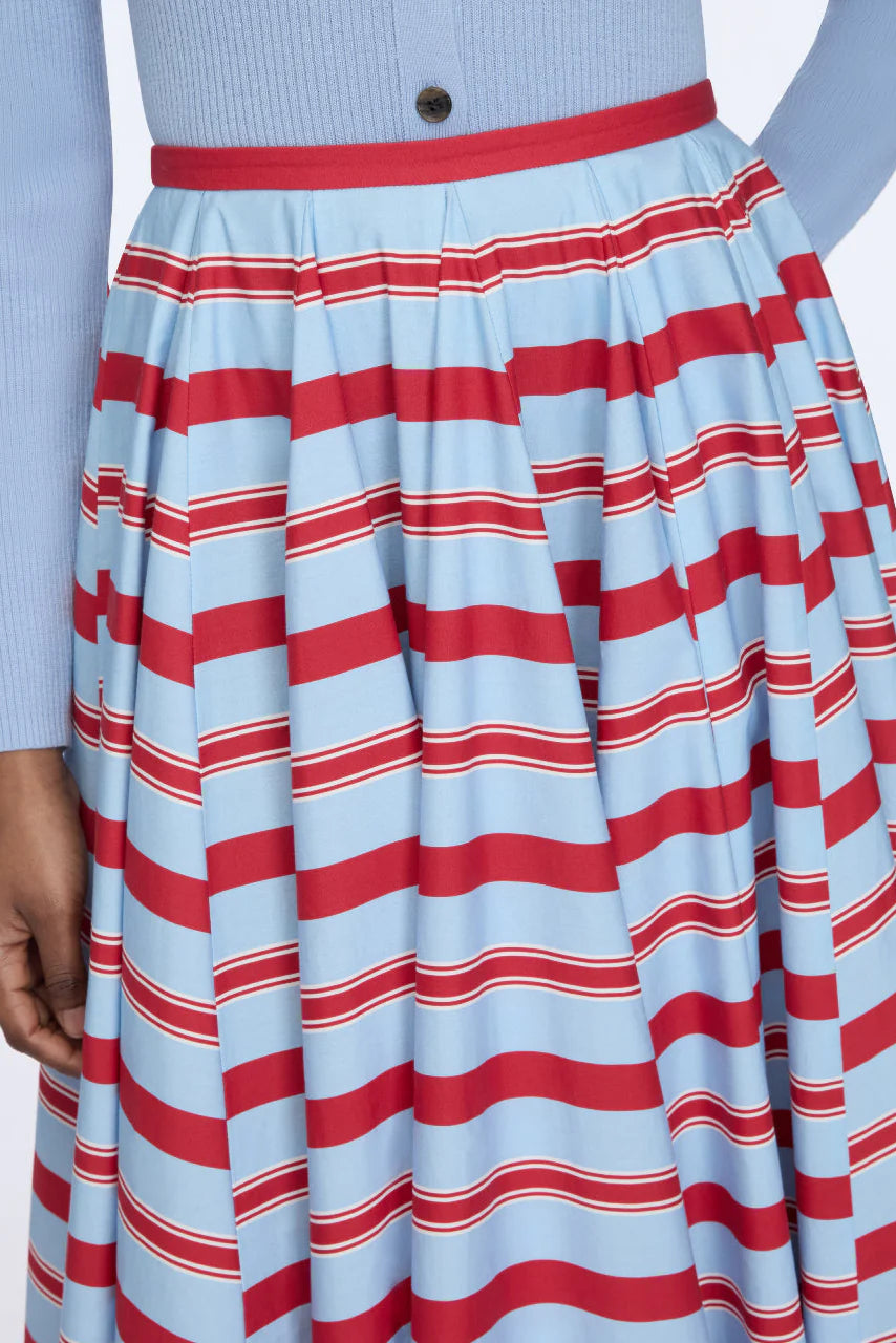 A person wearing a light blue ribbed top tucked into the Rhea Skirt by Emilia Wickstead, a full, pleated cotton skirt featuring horizontal red, white, and blue stripes.