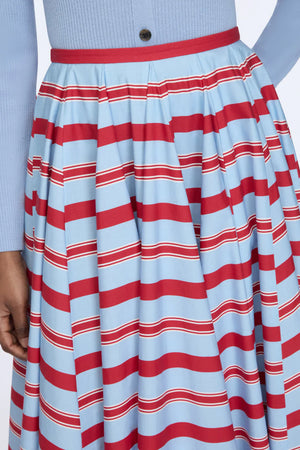 A person wearing a light blue ribbed top tucked into the Rhea Skirt by Emilia Wickstead, a full, pleated cotton skirt featuring horizontal red, white, and blue stripes.