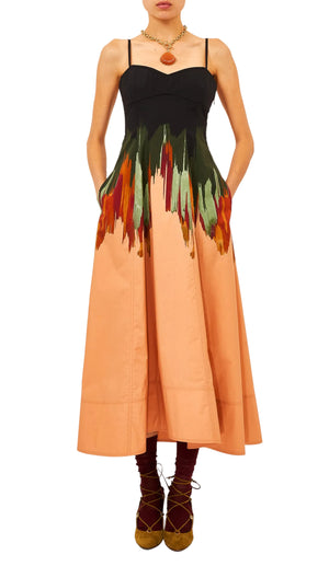 A woman wears the Fiora Cami Dress by Ulla Johnson, a sleeveless black and peach midi dress with a multicolored abstract brushstroke pattern, styled with brown lace-up shoes and layered necklaces.