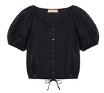 The Clara Top by Ulla Johnson is a black short-sleeve blouse featuring front buttons, pintuck details, delicate Victorian lace accents, a scoop neckline, puffed sleeves, and a drawstring hem.