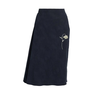 The Flower Jacquard Drape Midi Skirt by Erdem is a dark navy midi wrap skirt with subtle white floral embroidery on the side, inspired by tattoo-like motifs from Erdem’s Pre Fall 2026 collection.