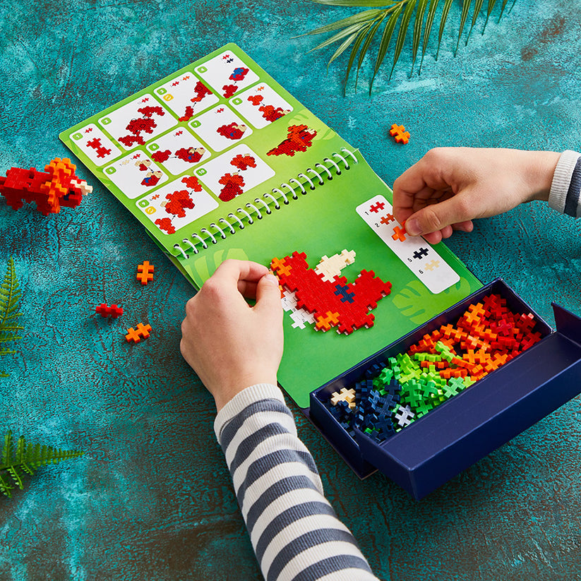 Using the Plus Plus Puzzle By Number® Activity Pad, a person assembles red, white, and orange pieces by following illustrated instructions in the spiral-bound activity pad on a blue-green tabletop—an engaging activity that sparks creativity and helps develop fine motor skills.