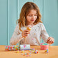 A young girl in a star-patterned shirt holds an ice cream cone while playing at a table with colorful pieces from the Plus Plus Gelato 240 Piece Tube, a pastel STEM building set.