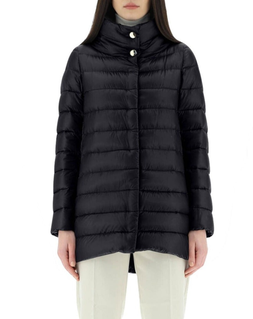 A person stands against a white background wearing the Herno Amelia Jacket, an ultralight nylon black puffer by Herno, featuring a high collar and white buttons. Paired with light pants and filled with cozy goose down, it effortlessly combines style and warmth.