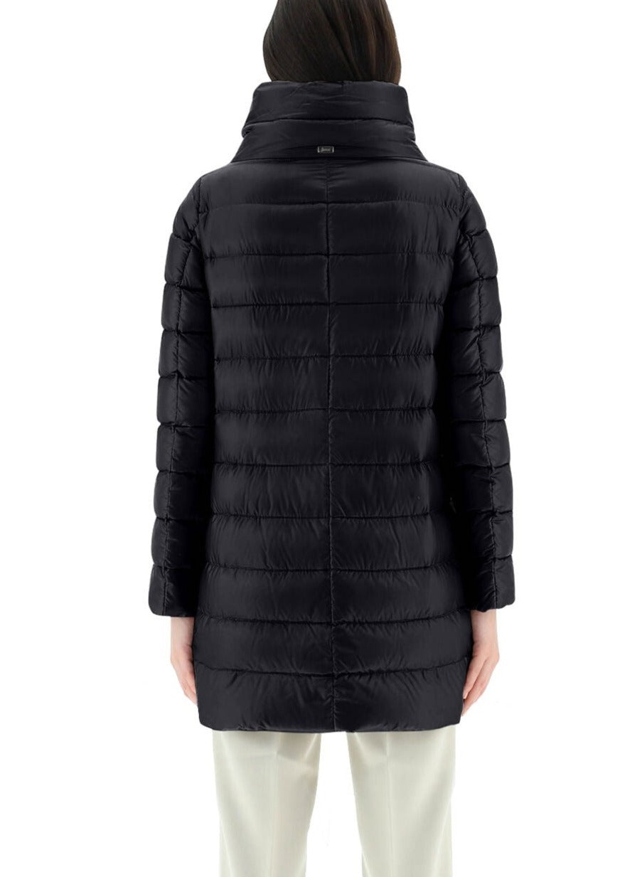 The individual is viewed from behind, donning a long, black Herno Amelia Jacket crafted from ultralight nylon and filled with goose down, complemented by light-colored pants against a white backdrop.