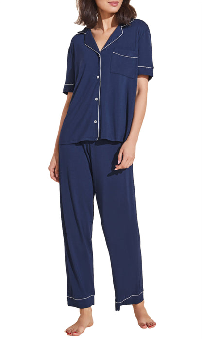 A woman wearing an Eberjey Gisele Short Sleeve Long Pant PJ Set with white piping, crafted from sustainable TENCEL™ Modal, featuring a short-sleeved button-up top and long pants, stands barefoot against a white background.