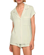 A person wearing the Eberjey Frida Shortie PJ Set: a light green slim fit short sleeve button-up top with a chest pocket, paired with matching low-rise cheeky shorts featuring an elastic waistband.