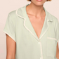 A woman, shown from the shoulders up, wears the Eberjey Frida Shortie PJ Set—a light green slim fit short sleeve pajama top with white trim and buttons.