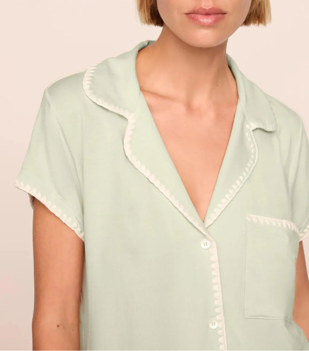A woman, shown from the shoulders up, wears the Eberjey Frida Shortie PJ Set—a light green slim fit short sleeve pajama top with white trim and buttons.